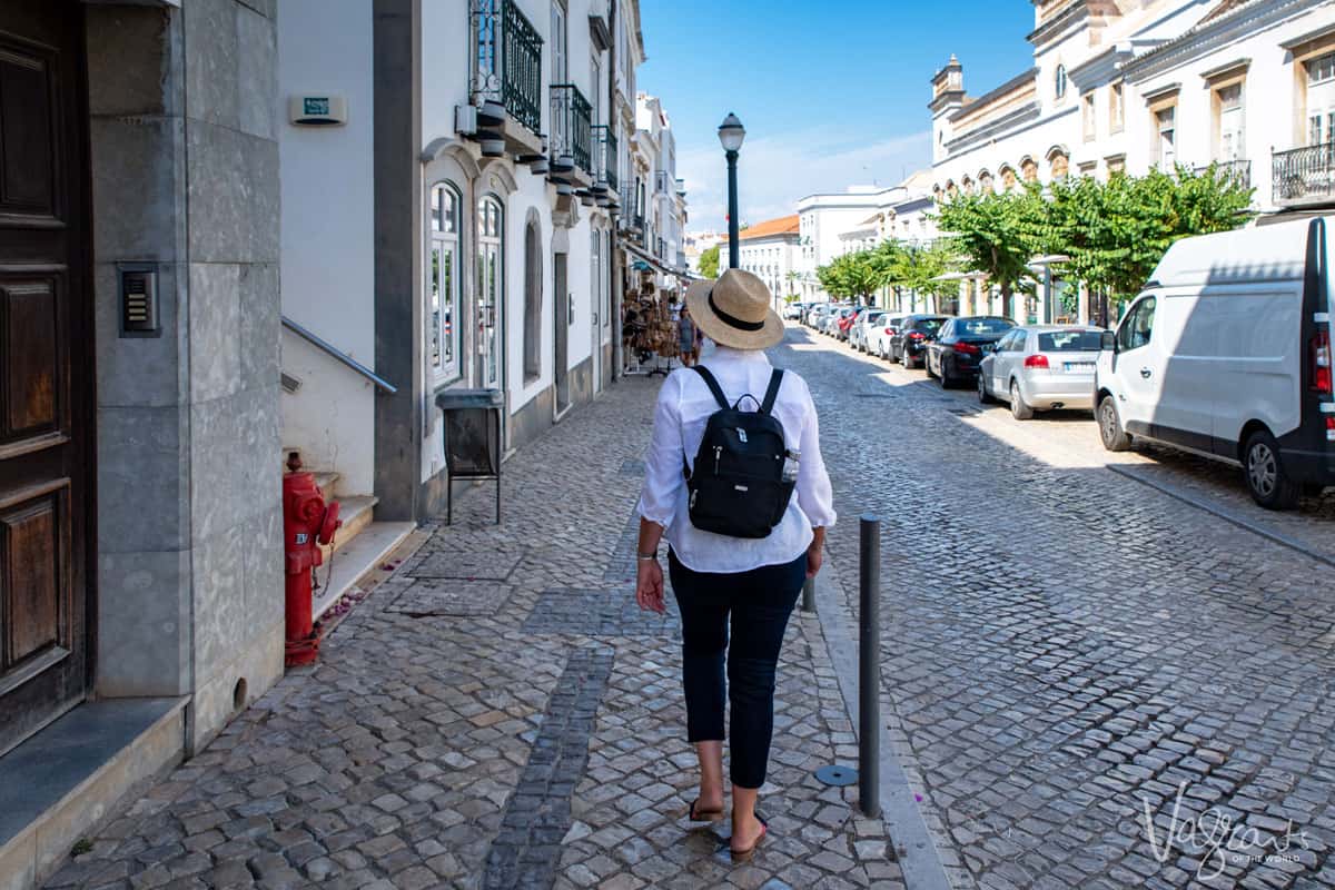 A woman in a white shirt and straw hat wearing a backpack wanders through the cobbled streets of old town Tavira. 
