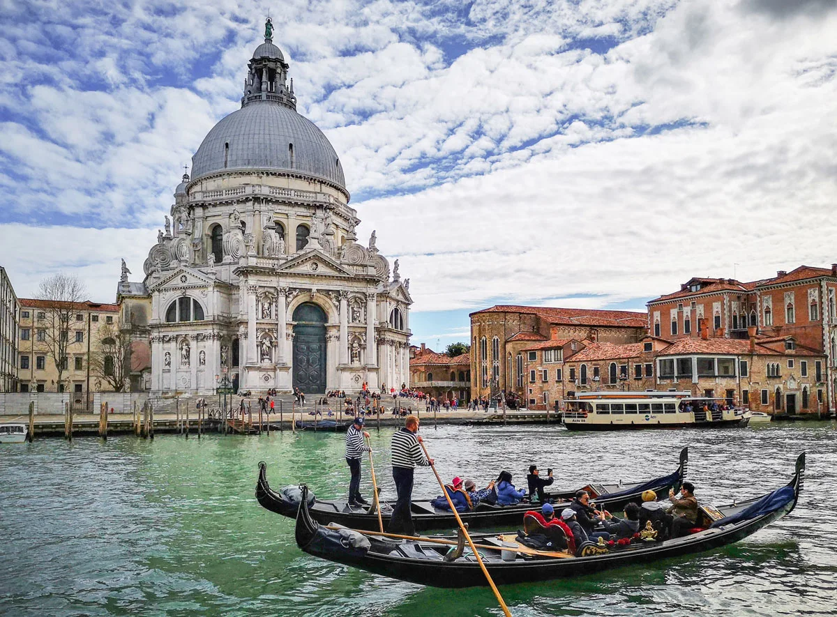 Two traditional gondola boats carry tourists down the Venice lagoon in front of a historical domed church building.