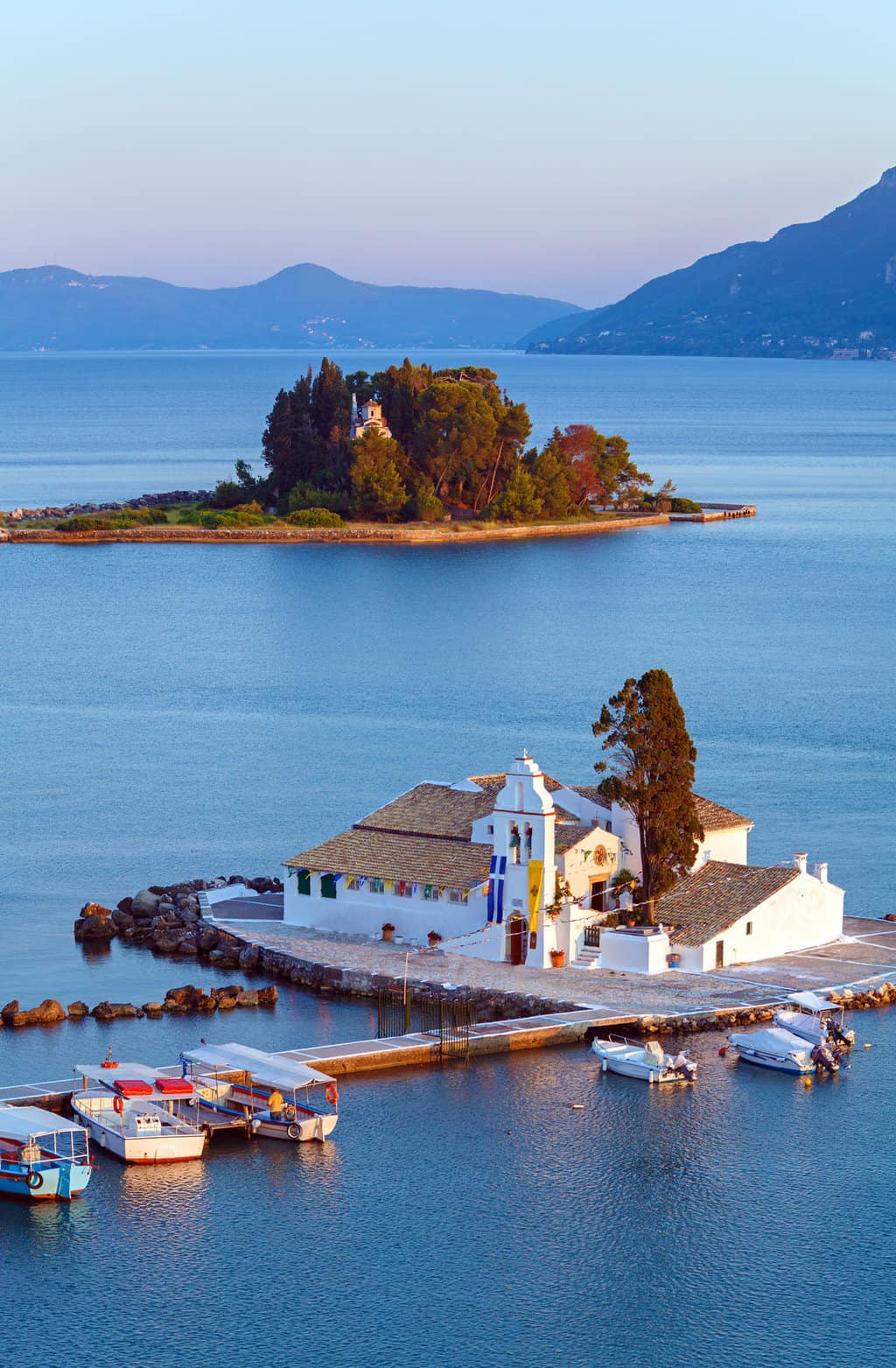 Two small islands with typical Greek churches sit in the middle of calm, blue ocean. Boats are moored in front of the closest island. 