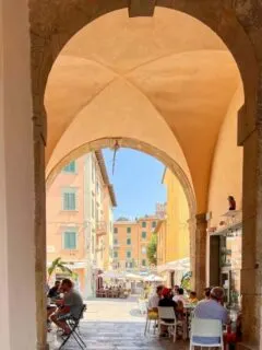 A stone arched walkway lined with cafe tables leading to a plaza in the old town of Elba Island.