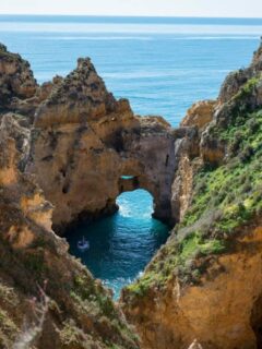 Yellow cliffs covered in bright green plants form a circular open cave around a tourist boat in clear calm water on the coast of Lagos Portugal.