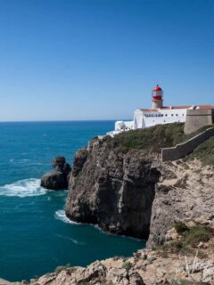 The red and white Cabo São Vincente lighthouse sits on the top of sheer cliffs that fall into a calm blue sea in Sagresh Portugal. It is a clear, sunny day.