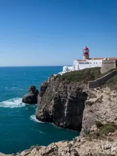 The red and white Cabo São Vincente lighthouse sits on the top of sheer cliffs that fall into a calm blue sea in Sagresh Portugal. It is a clear, sunny day.