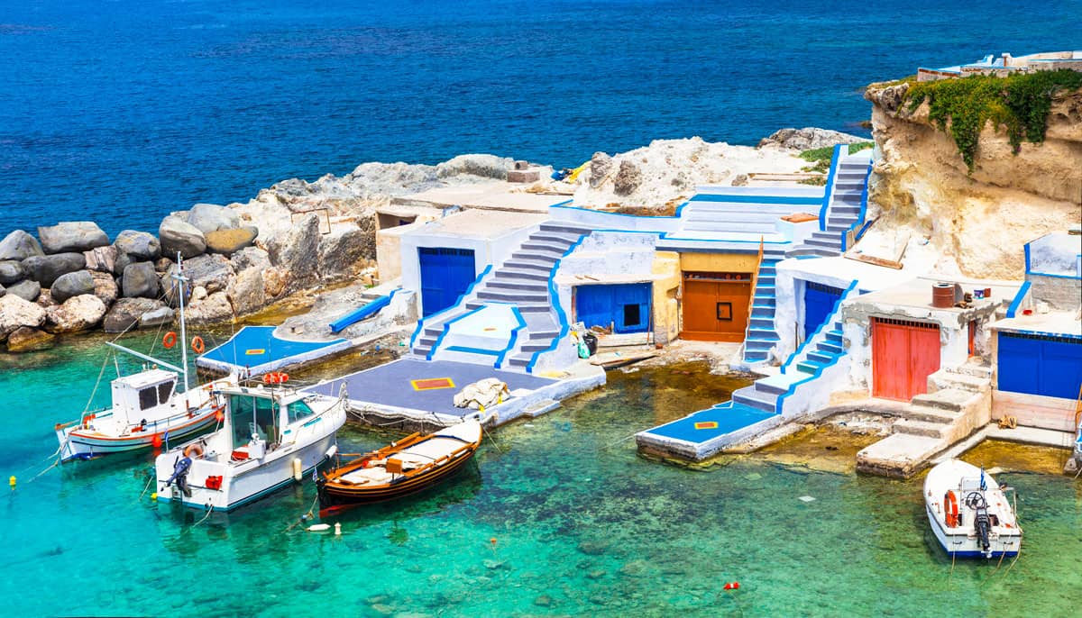 Colourful fishing boats moored in front of brightly painted boat shed with colourful stairs leading from the docks on Milos Island Greece. 