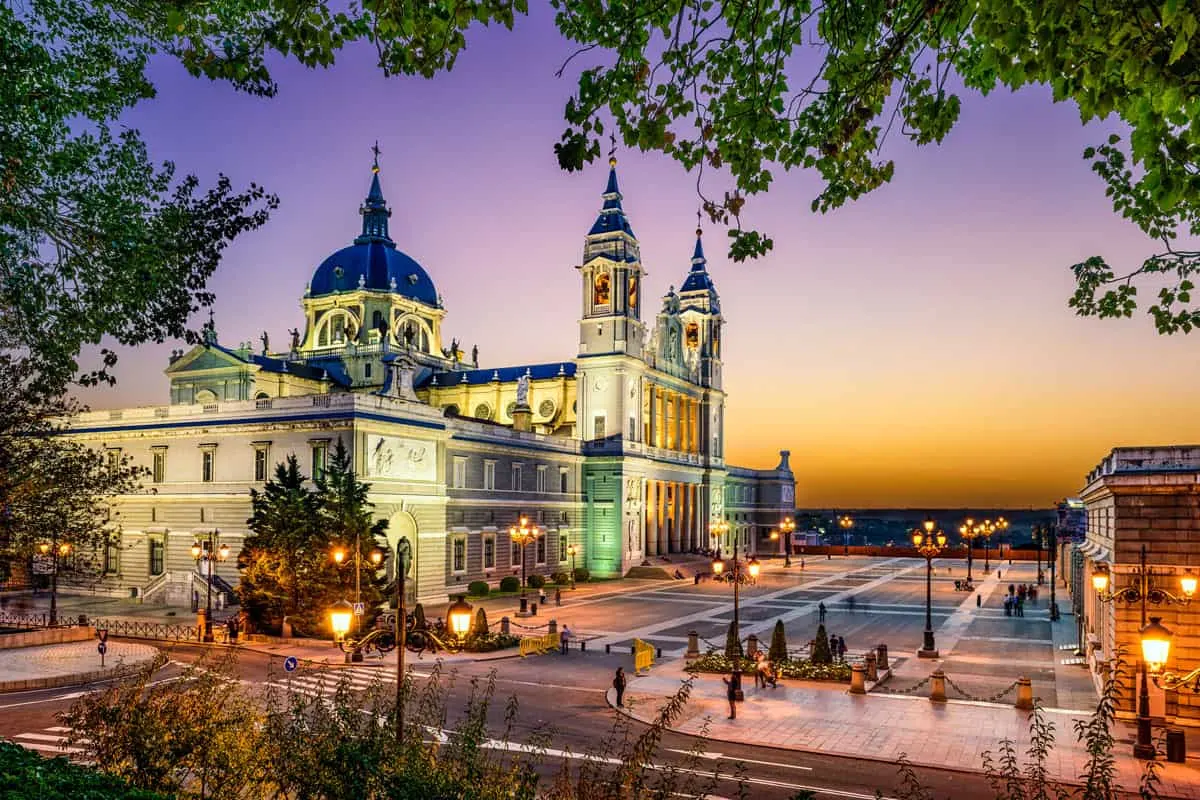 The Almudena cathedral at dusk with a pink and orange sky. The building is lit with soft lighting emphasising the spires and domes. 