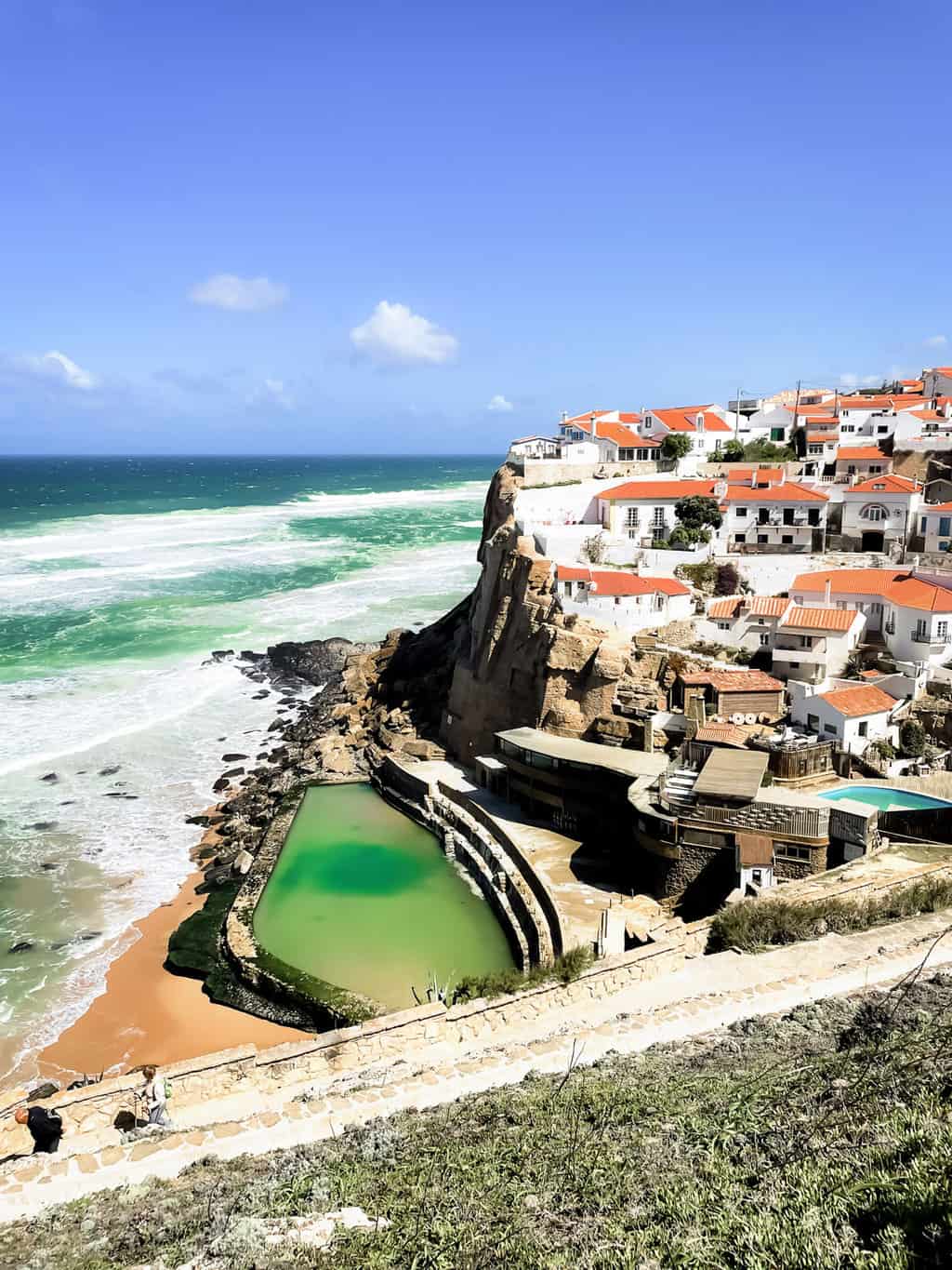A small village with white houses and red roofs overlook the sea and a man made ocean pool in Azenhas do Mar Portugal. 