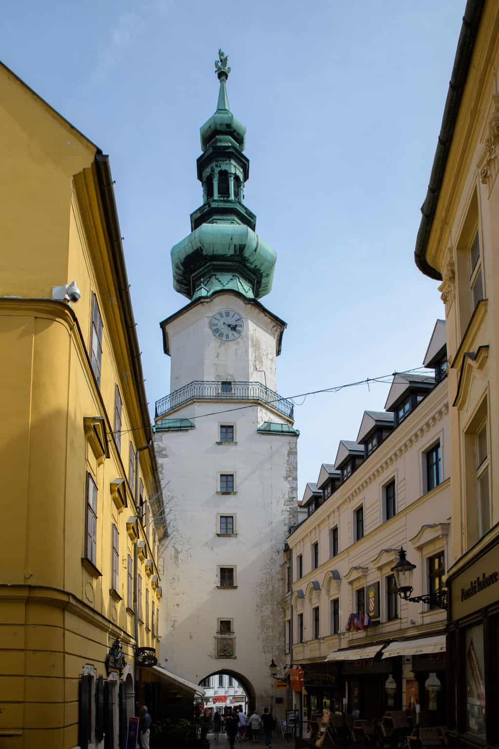 A narrow street in Bratislava with yellow buidling wiether side and an onion domed spire over an archway. 