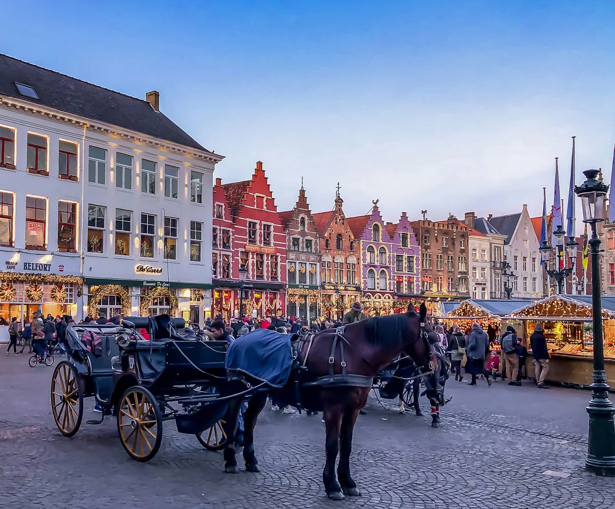 A horse drawn carriage in front of a twinkling christmas market and the colourful gabled houses in Bruges. 