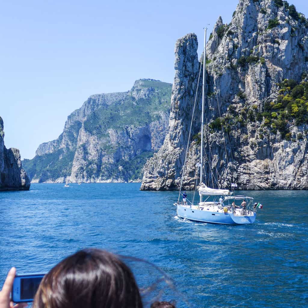 A blue yacht sails past rugged cliffs on the island of Capri in Italy on a sunny day.
