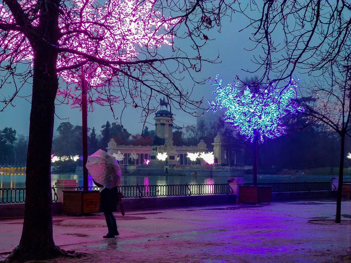 Christmas lights decorating the Retiro Park in Madrid. A man stands next to the pond with an umbrella on a gray and rainy day, in Spain.