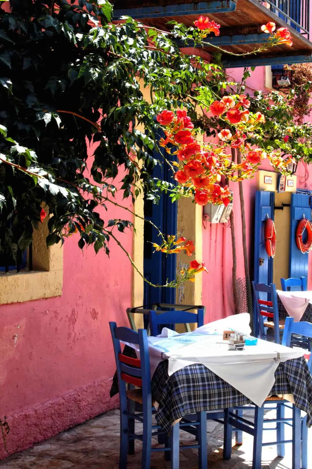 Typical Greek restaurant table outside a bright pink building with pink flowers hanging over the wall.