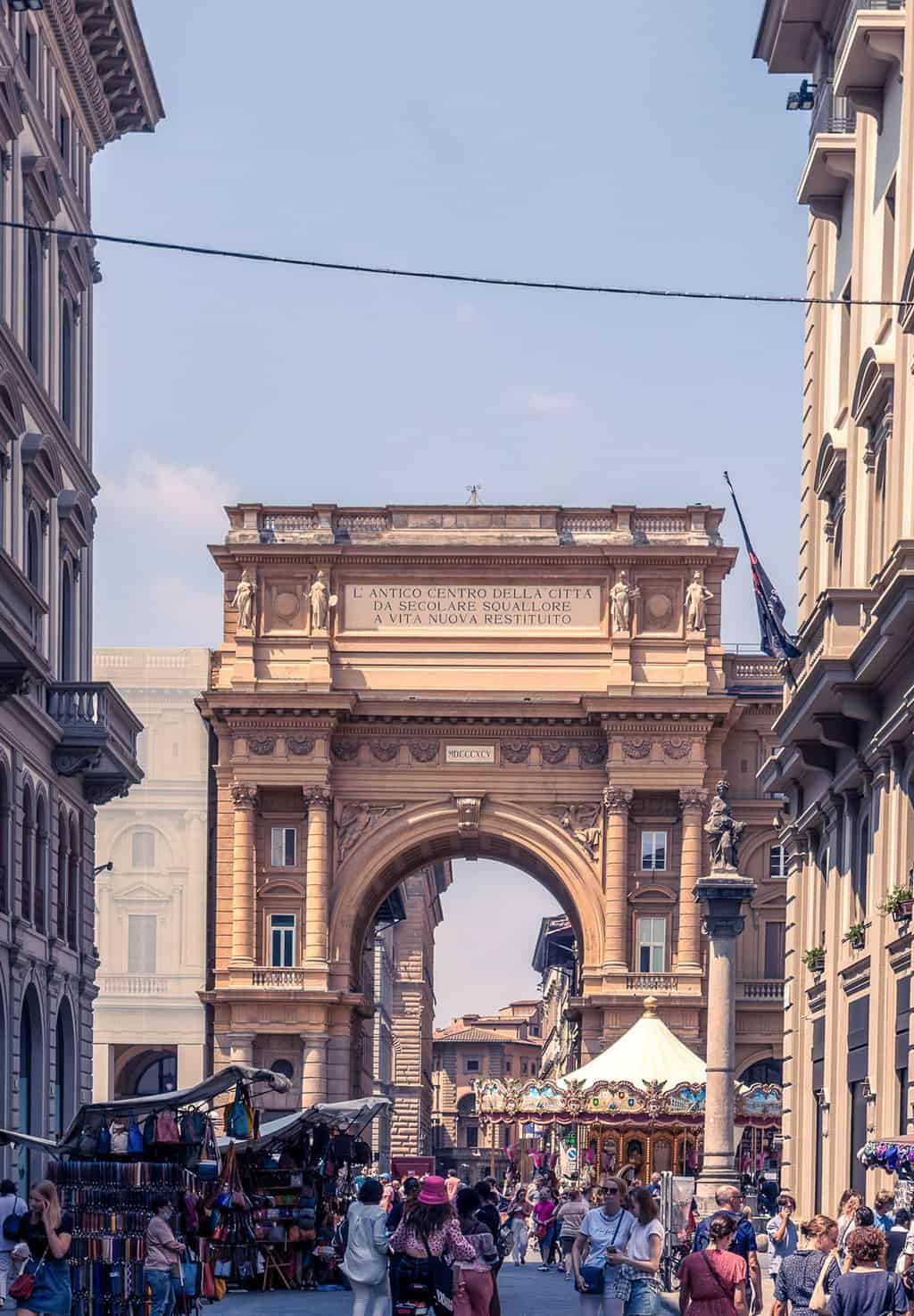 Tourists walking in Florence. there is a large arch at the end of the street and an old fashioned carousel. 