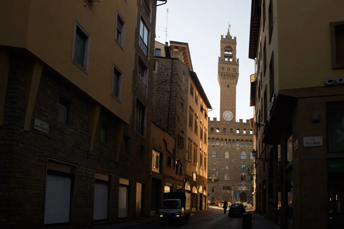 The clock tower of the Palazzo Vecchio seen at the end of a narrow street in the orange morning light. 