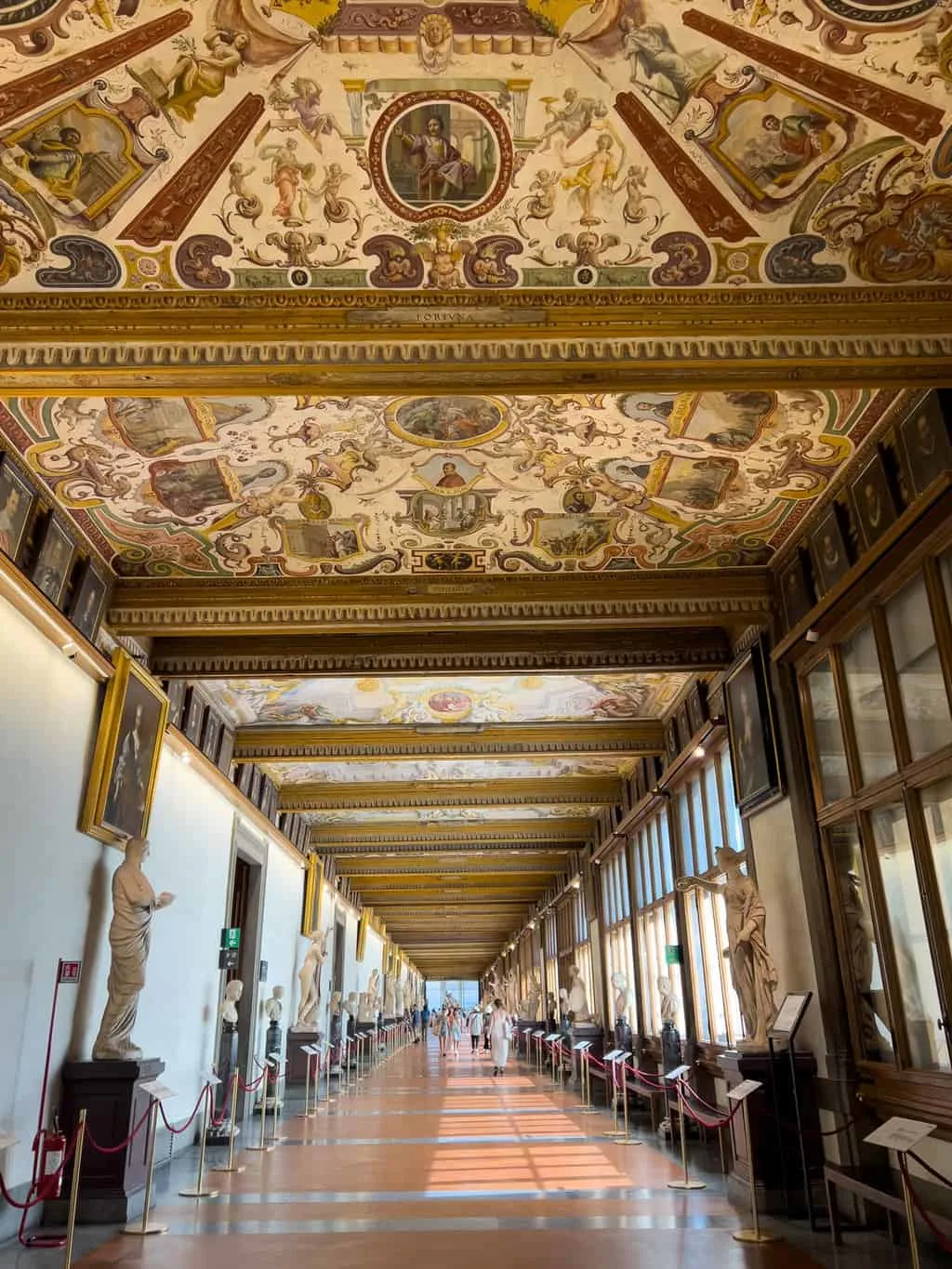 A long corridor in the Uffizi Museum in Florence. There are frescos painted on the panneled ceiling with statues lining the brightly lit corridor. 