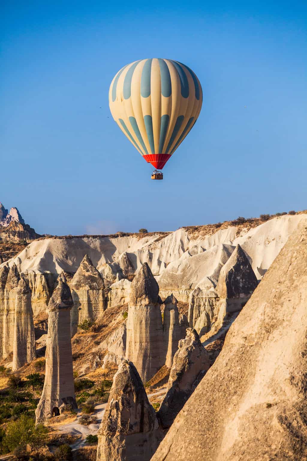 A hot air balloon flies over the fairy chimney rock formations in Cappadocia Turkey on a clear morning. 