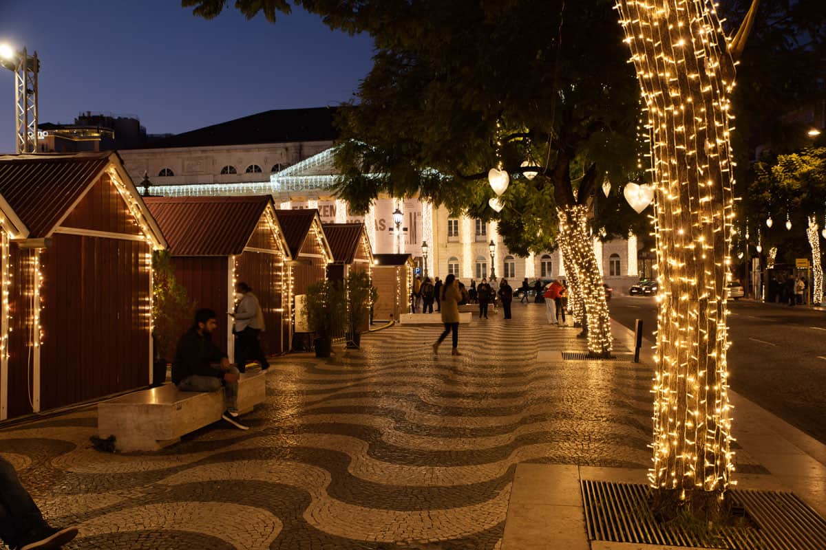 Christmas market huts and Christmas lights at night in Lisbon. people are walking across the typical black and white mosaic pavements of Portugal. 