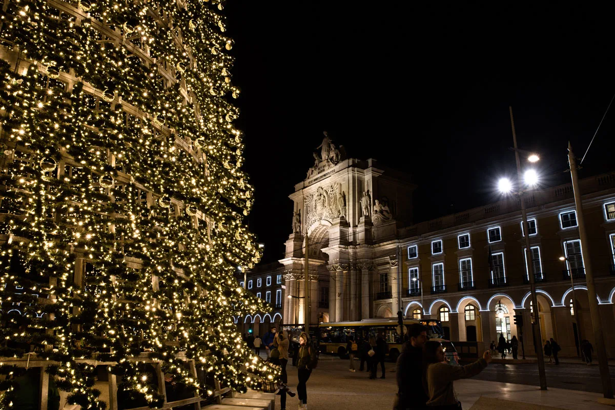 A girl standing in front of an enourmous Christmas tree in the main square of Lisbon at night. 