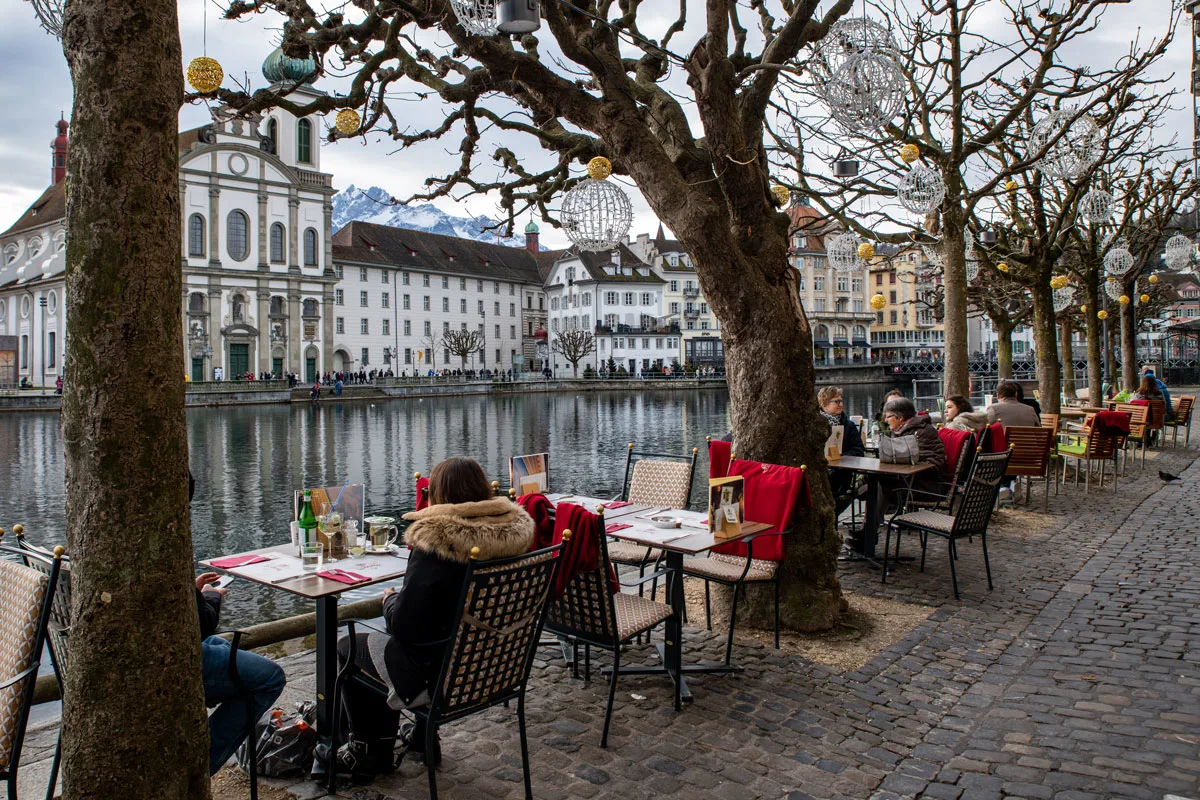 People sitting at an outdoor cafe by the river in the beautiful city of Lucerne in Switzerland. It is winter and there are red blankets on the cafe chairs. 
