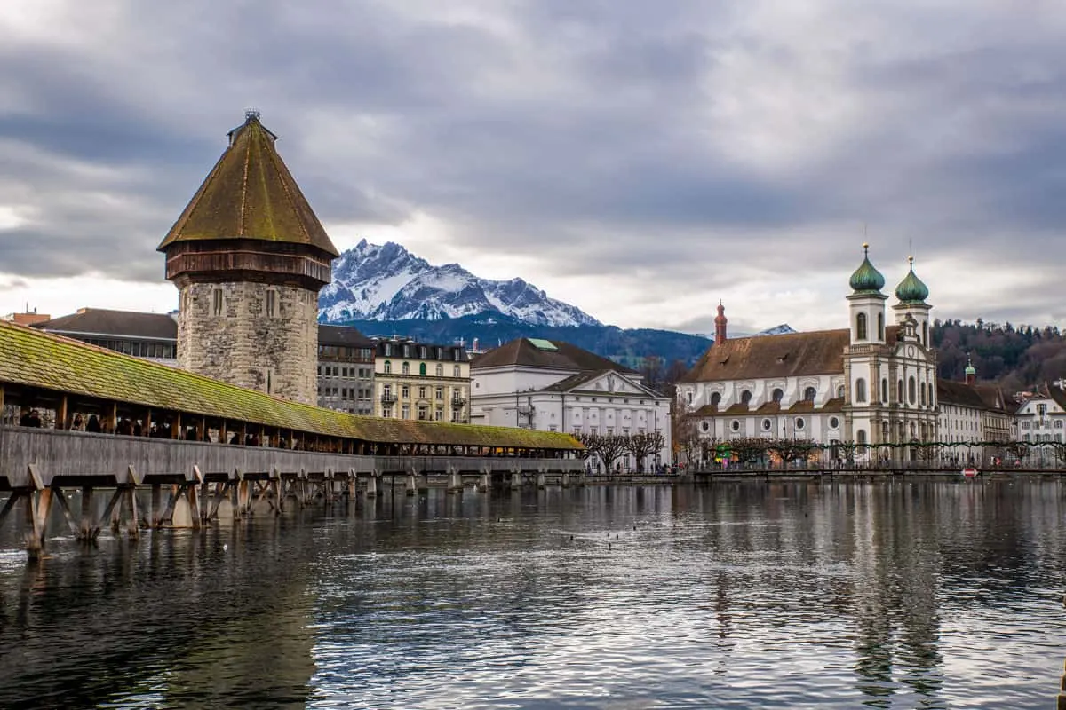 Snow capped mountains in the distance behind the iconic covered bridge on the river in Lucerne Switzerland. The double, onion dome spires of the church create a pretty skyline next to the mountain. 
