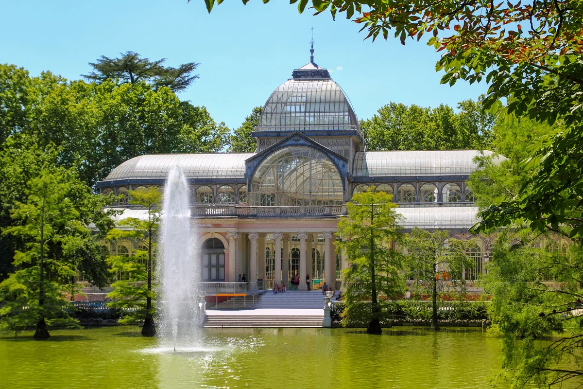 A glass and steel atrium style building in front of a lake with a fountain in Retiro Park Madrid. 