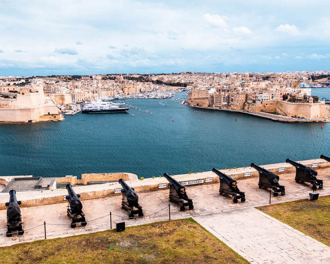 Cannons lined up overlooking the harbour and two cities of Malta. There are large boats moored in the harbour.