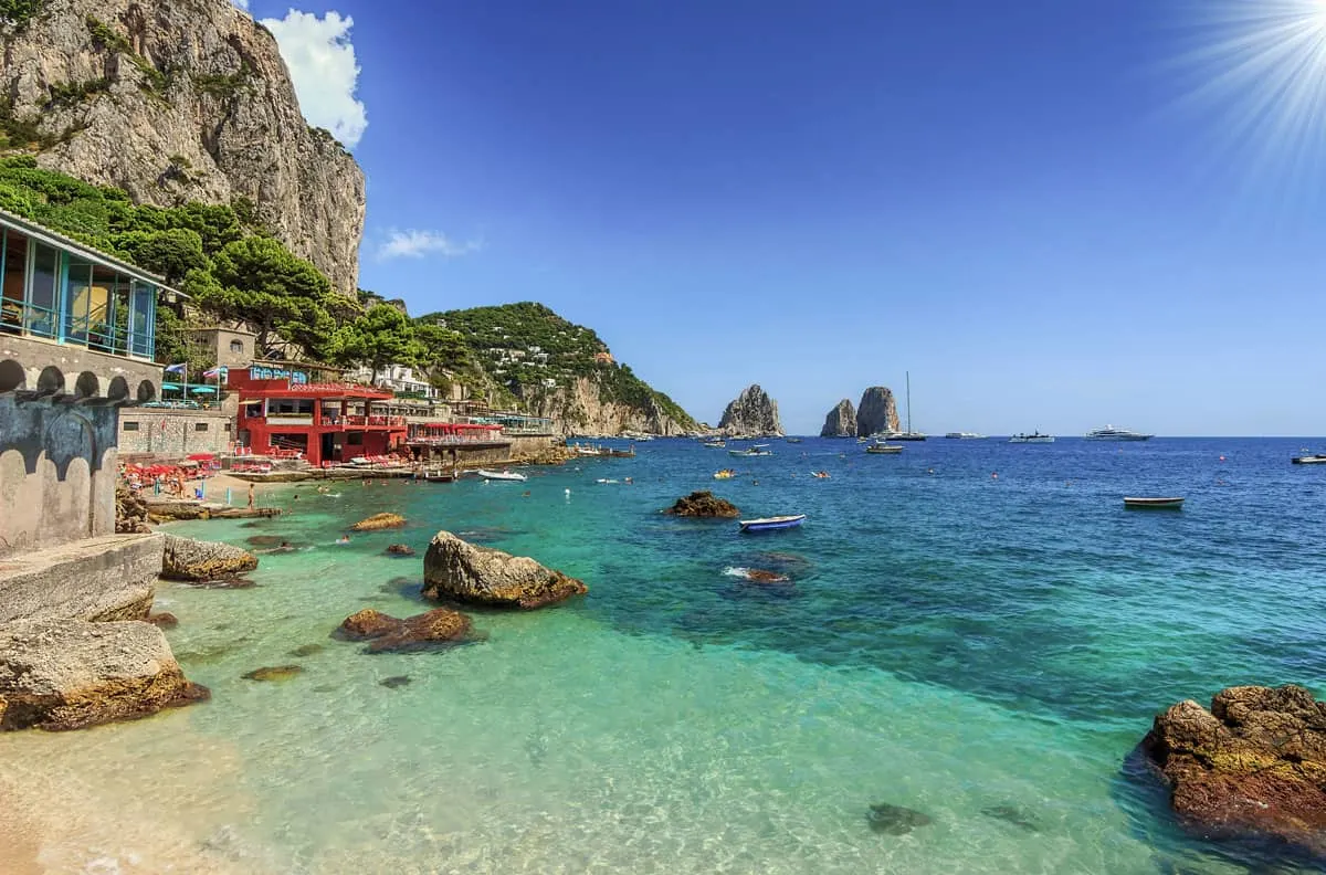 Clear water on a beach with rugged cliffs in the background and colourful beach bars overlooking the water.