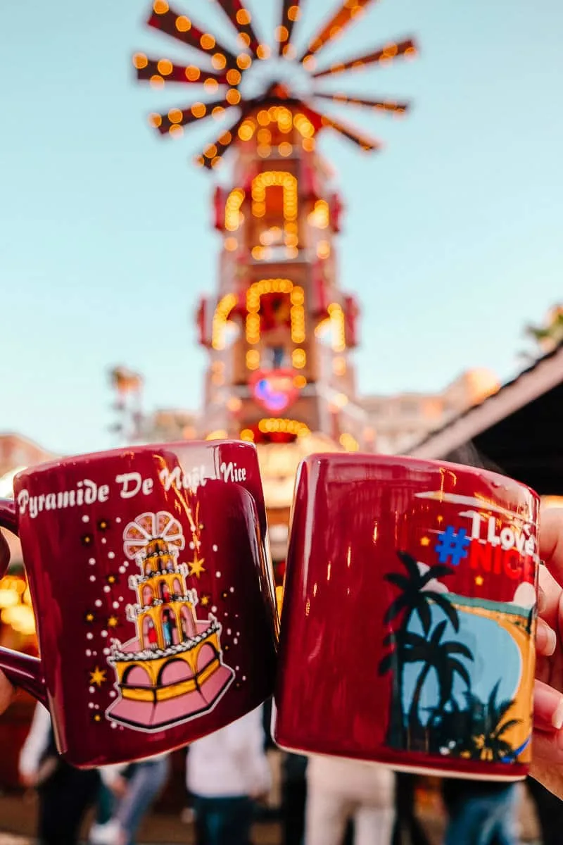 Two coffee mugs with mulled wine being clinked together with festive lights in the background in Nice in December.