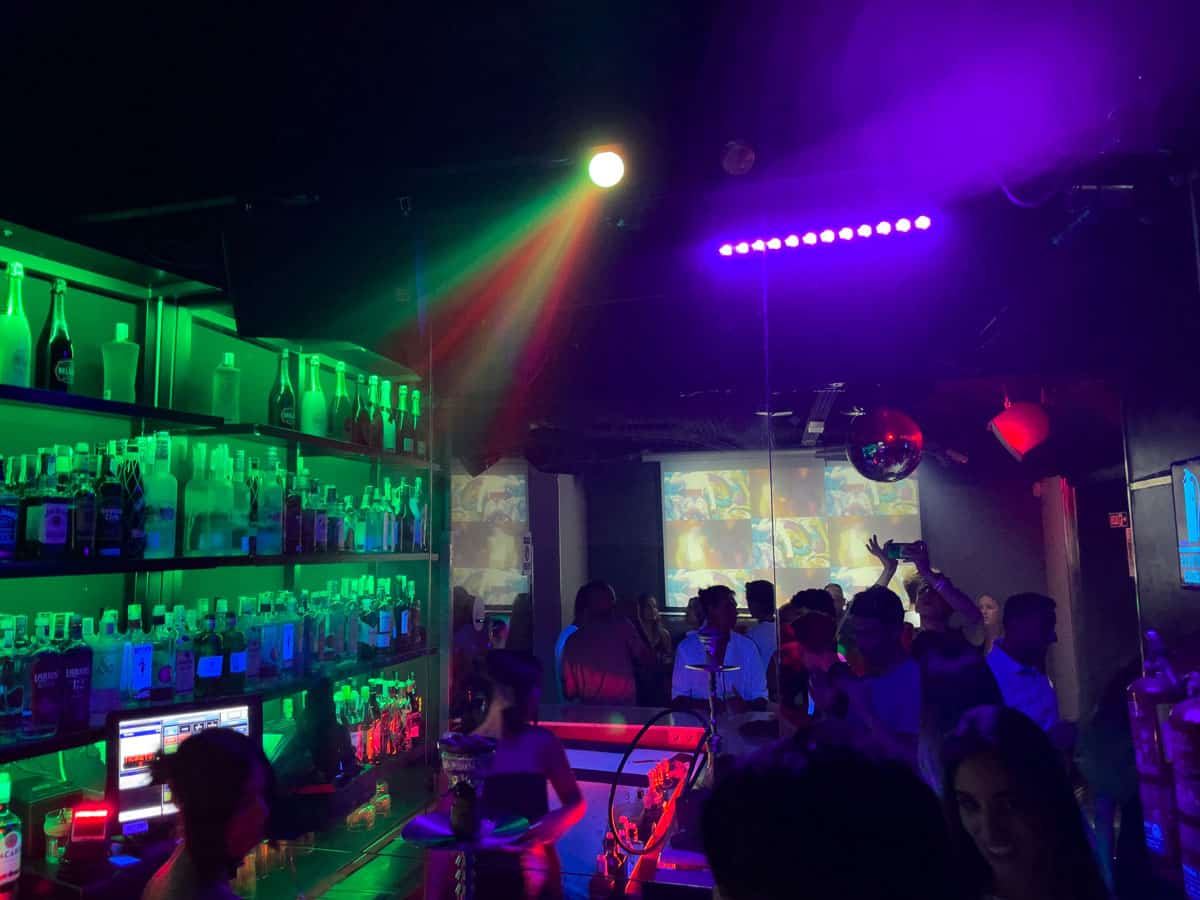 People in a dark nightclub with coloured lights reflecting off the bar. 