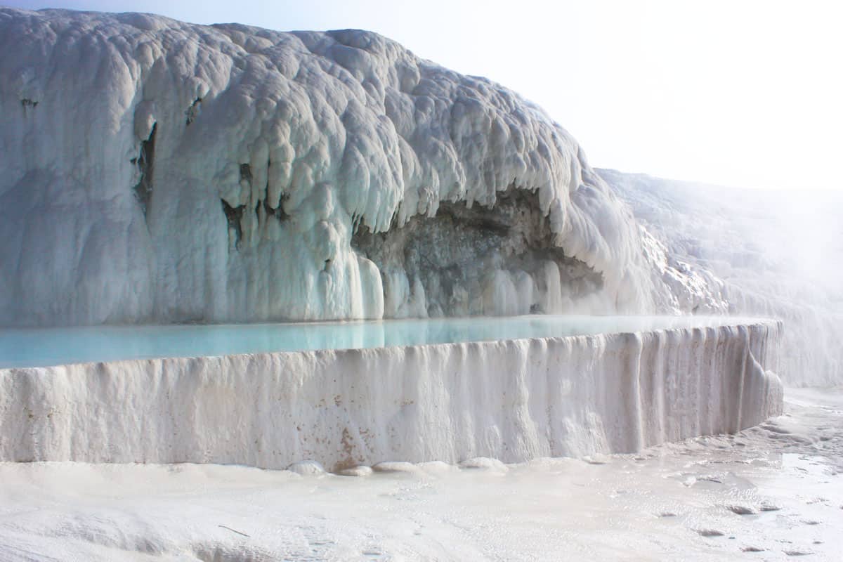 White travertine deposits cover rocks while steam rises from a clear blue natural spa. 