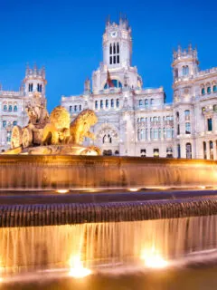 A large flowing fountain with lions and chariots in front of a palace lit up at night in Madrid.