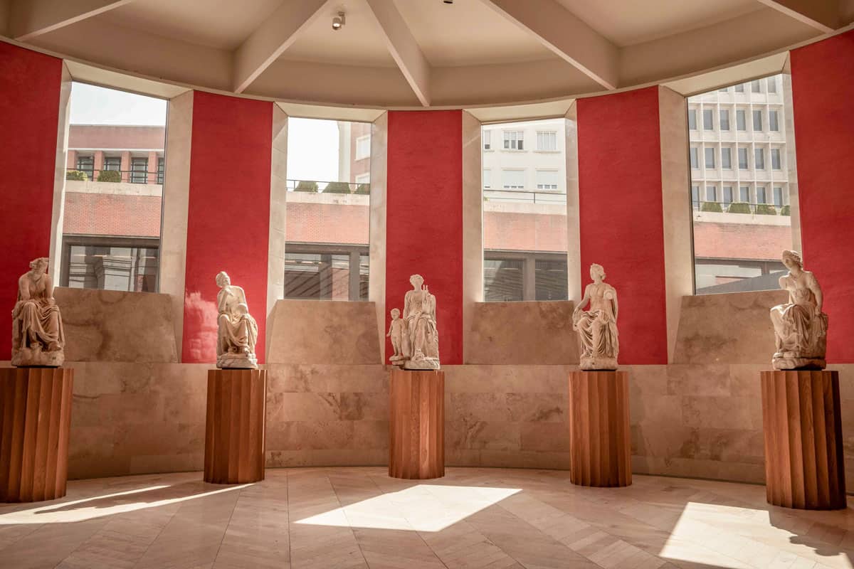Statues on wooden pillars against a red wall with windows in the Prado Museum in Madrid. 