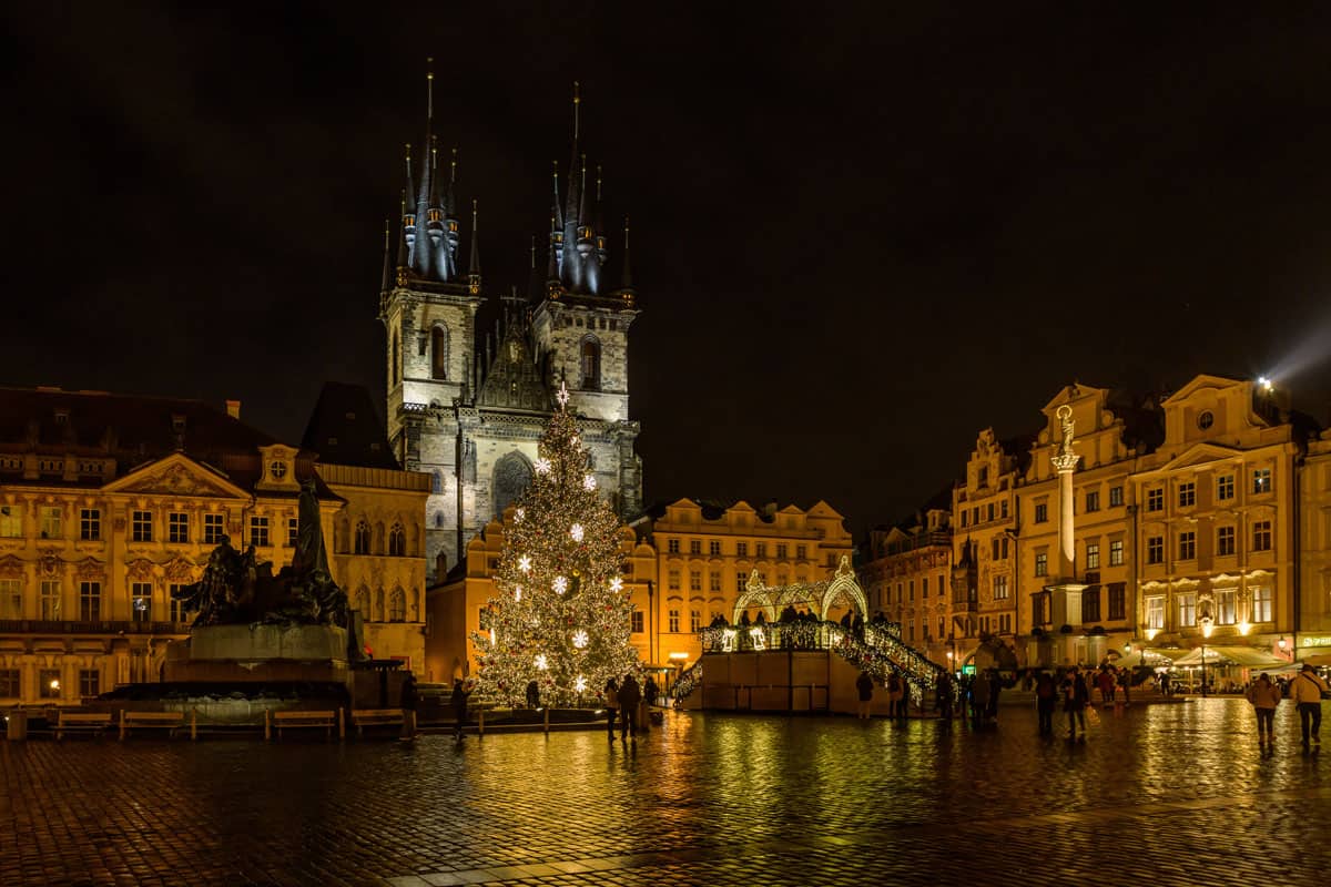 Night time in the main square in Prague. There is a large Christmas tree lit up in front of the iconic castle building with fairytale like spires on the roof. 