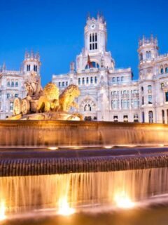 A large flowing fountain with lions and chariots in front of a palace lit up at night in Madrid.