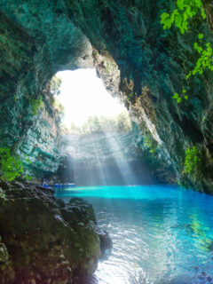 Light shines through a hole in the roof of a cave onto clear blue water. Row boats are in the light.