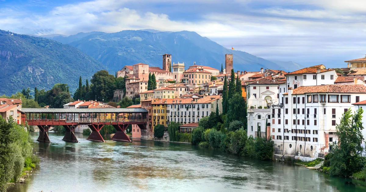 A covered bridge reflects in the river in a small Italian town with mountains in the distance. 