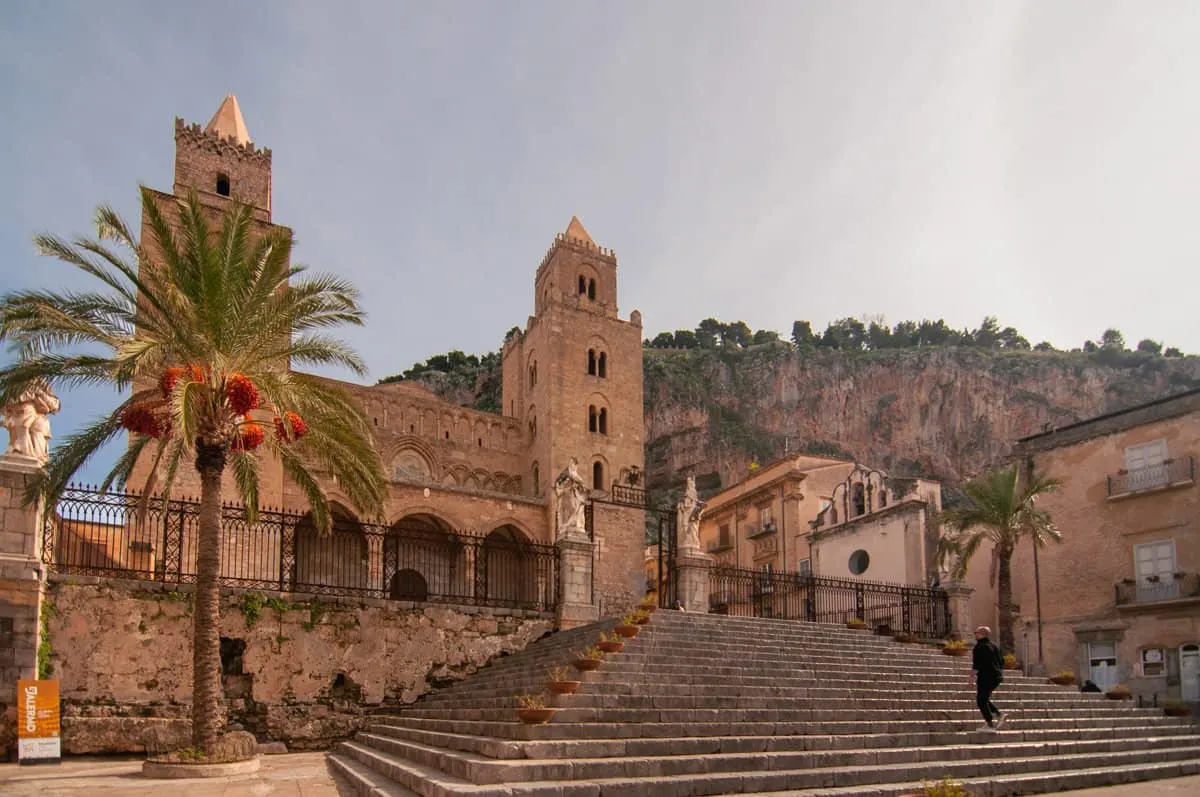 A man walks up the stairs to a stone historical cathedral in golden hues with a palm tree out the front.  
