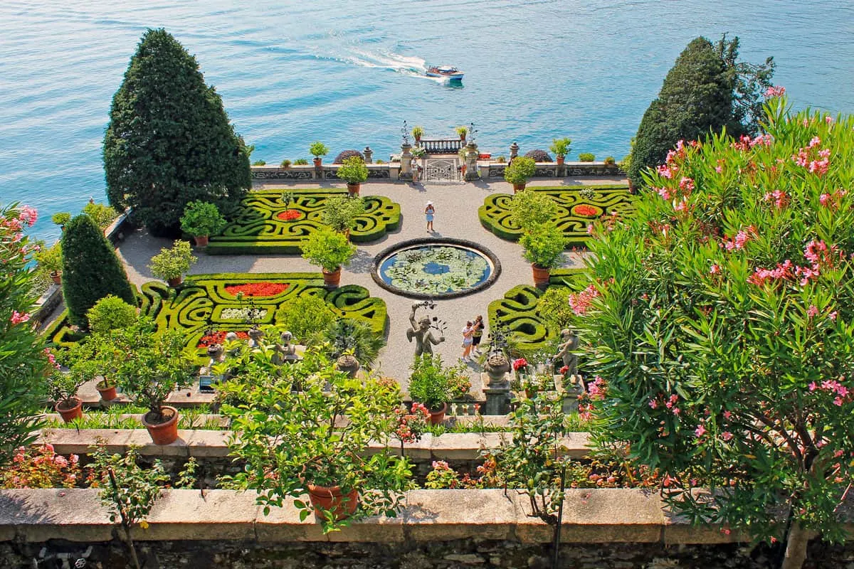 Ornate sculptured gardens overlooking a lake. A boat passes the gardens. 