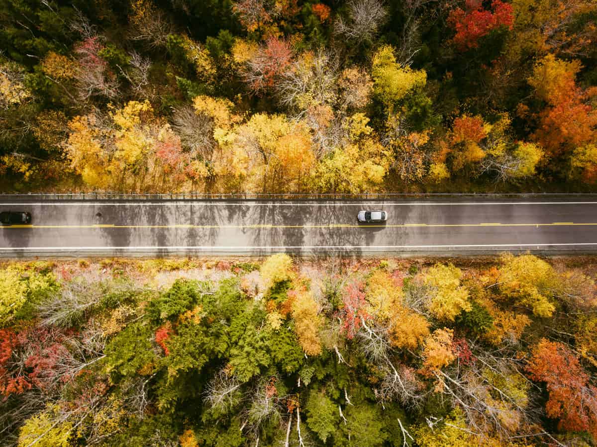 Arial view of a car driving down the Kancamagus Highway with dense fall foliage on either side of the road.