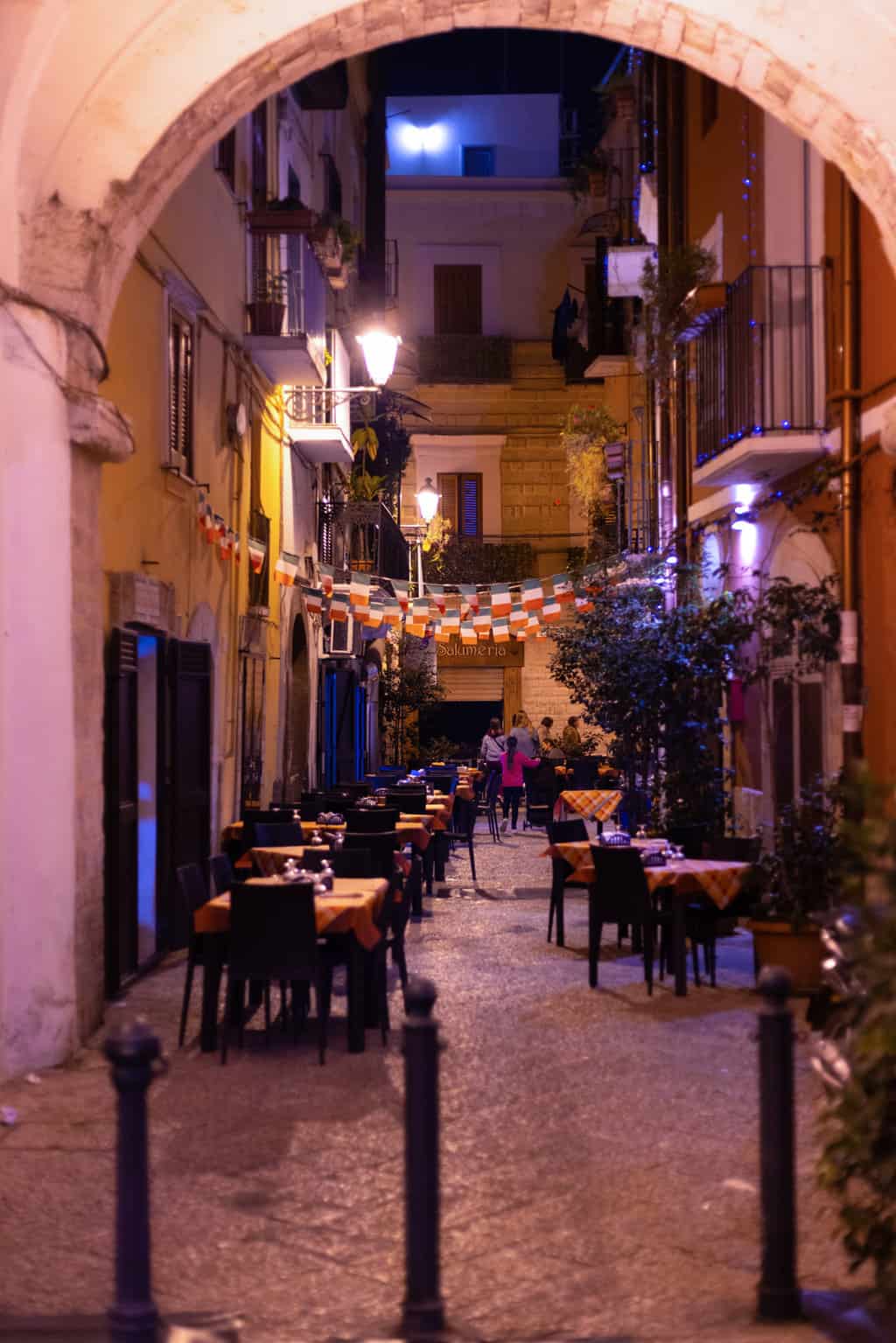 A narrow lane in Bari Italy at night with restaurant tables lining the street.