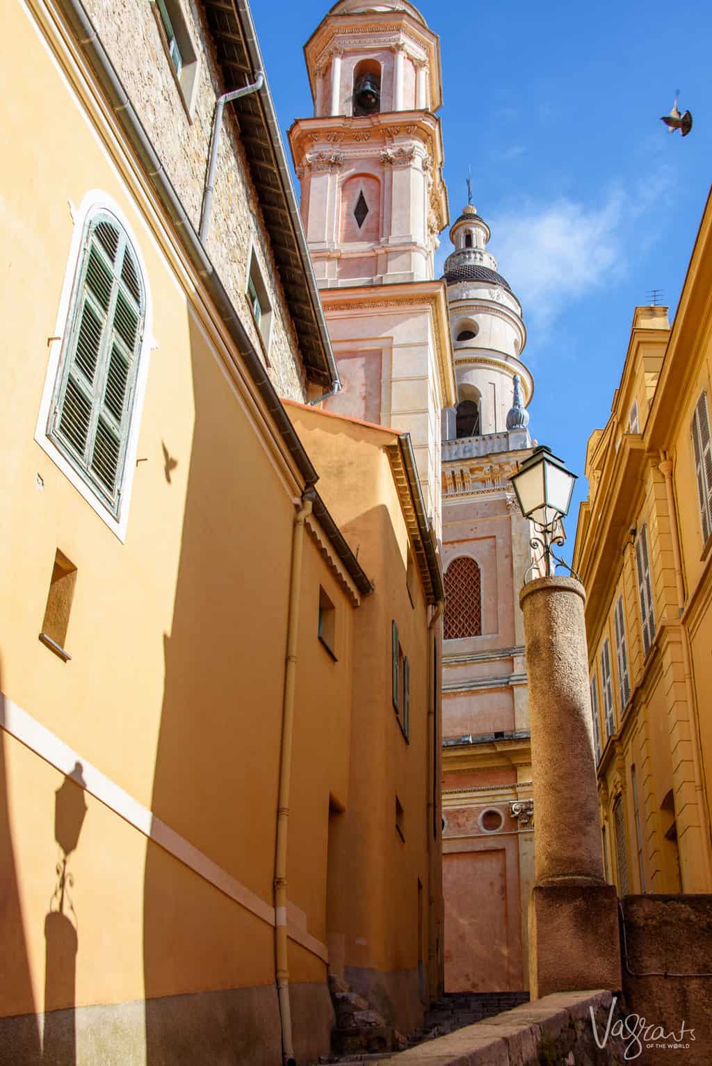 Looking down a narrow streets with neo classical buildings in hues of yellow and pink to the bell towers of a church. 
