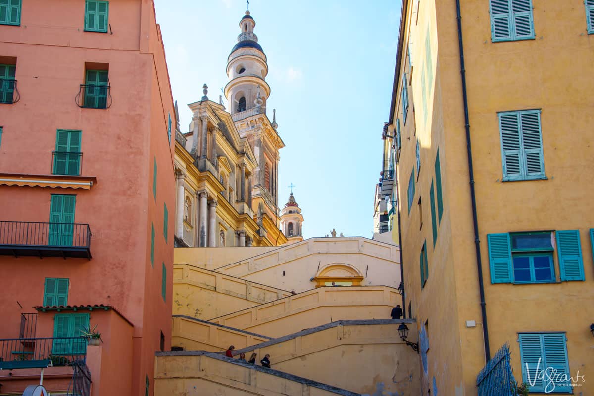Pastel coloured building either side of stairs leading to a church with a bell tower.