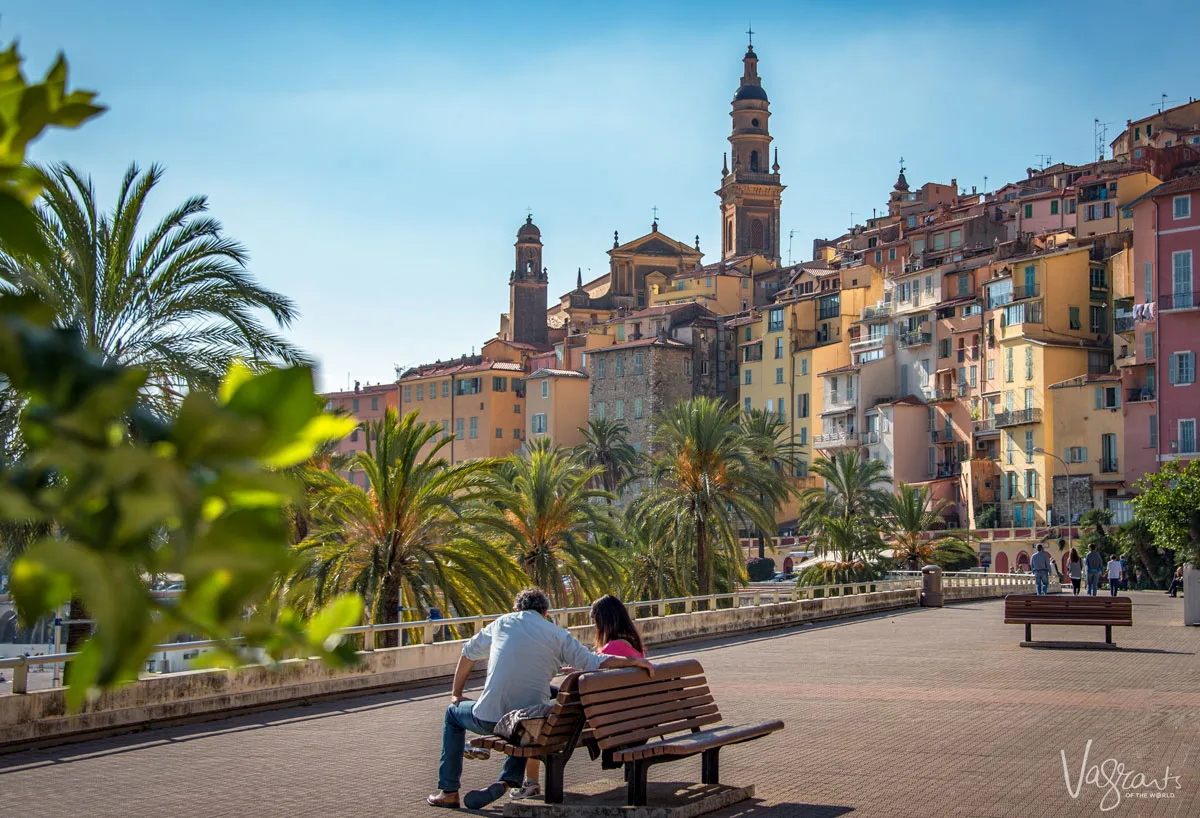 A couple sit on a park bench on a palm fringed promenade with the pastel coloured Menton old town in the distance. 