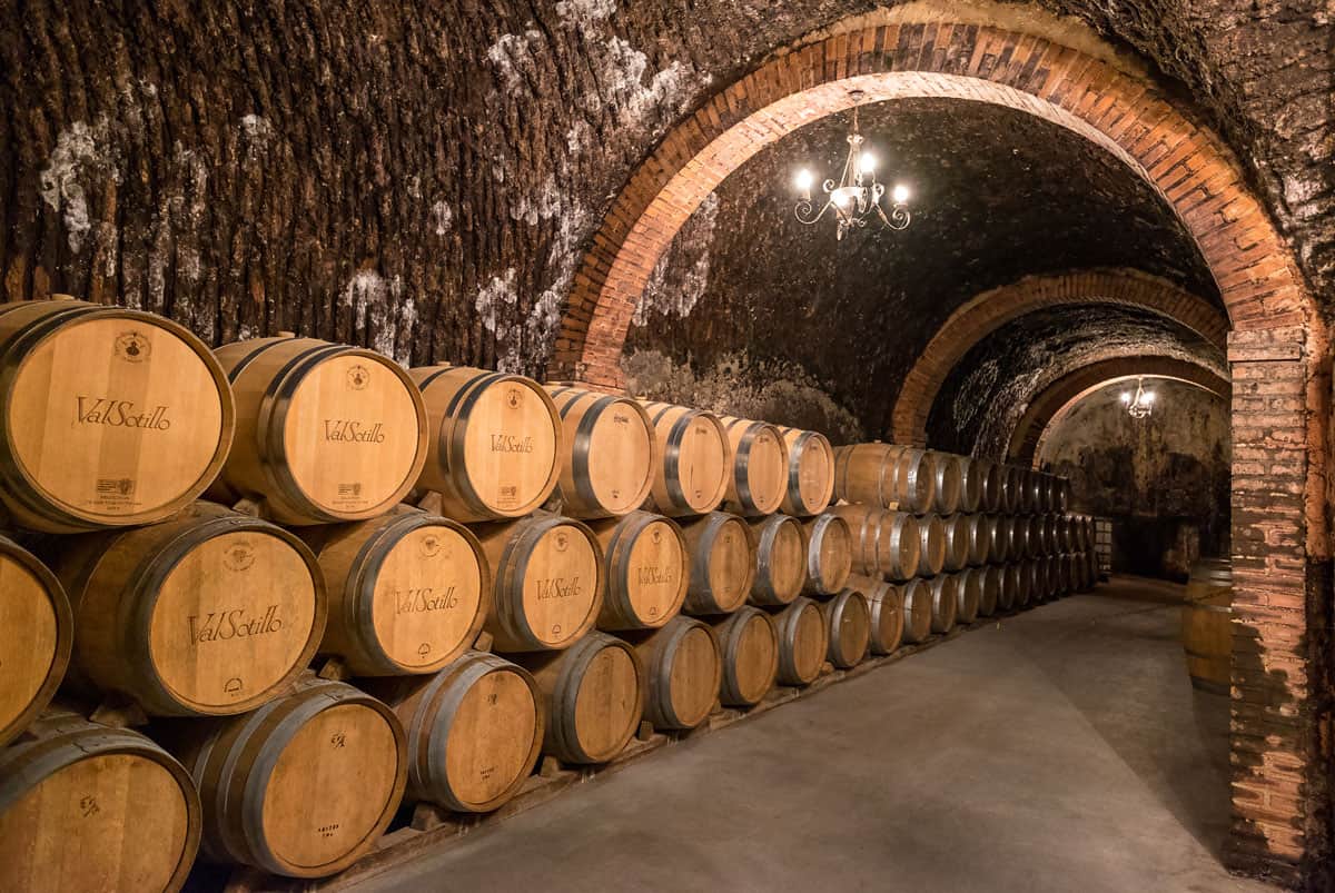 Oak wine barrels staked along a wall in a stone tunnel at a winery in Spain.