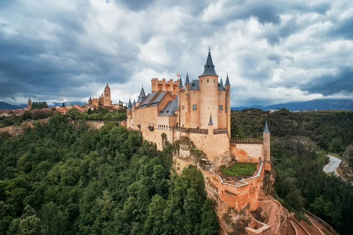 An historic castle that looks like a fairytale castle sits framed by trees on a rocky outcrop with storm clouds in the sky. 