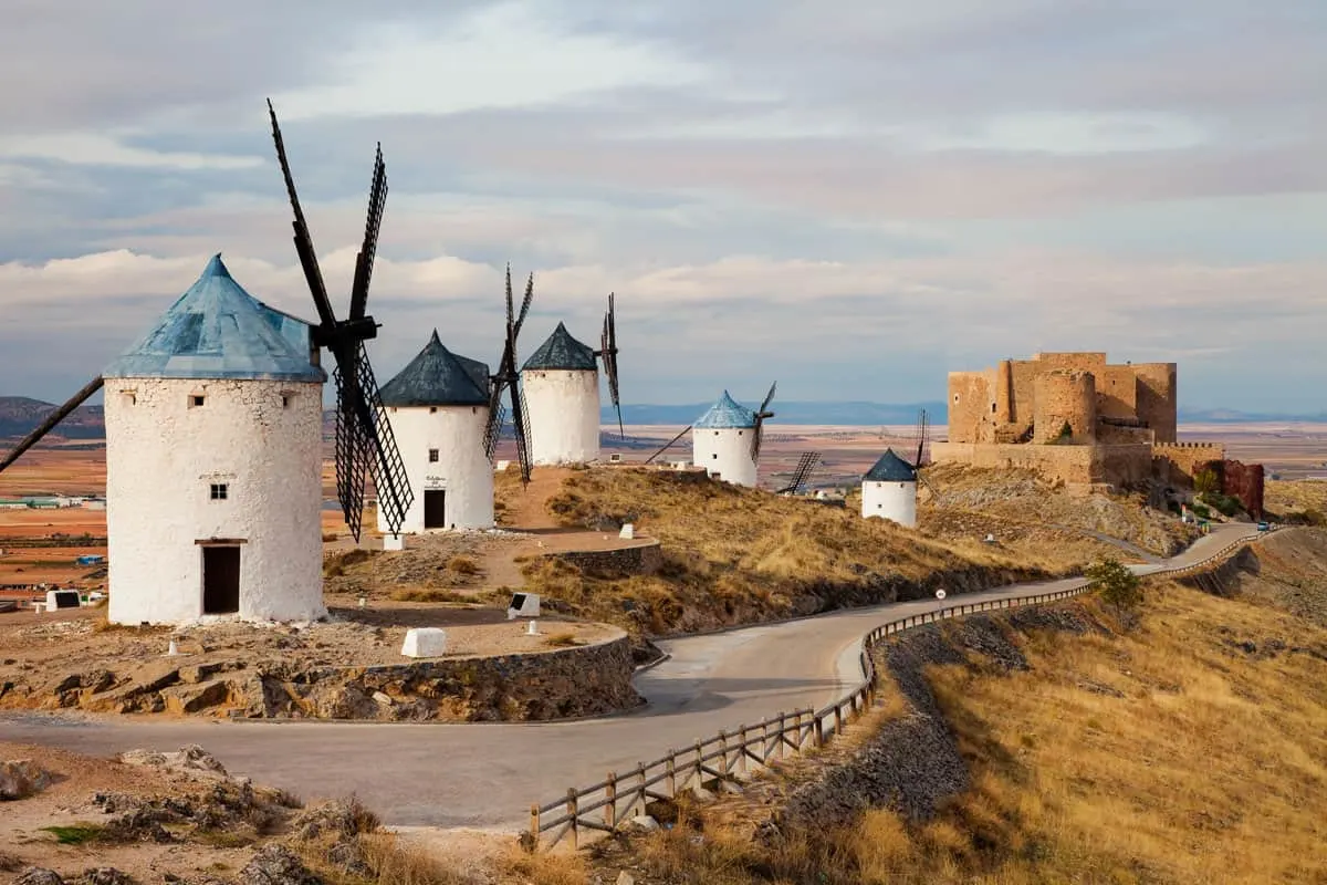 A row of traditional windmills in Spain surrounded by arid land. A ruin of a castle is in the distance. 
