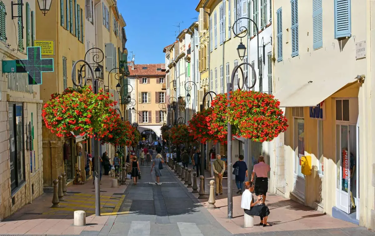 people walking down a pretty shopping street lined with flowers in Antibes on a summers day. 