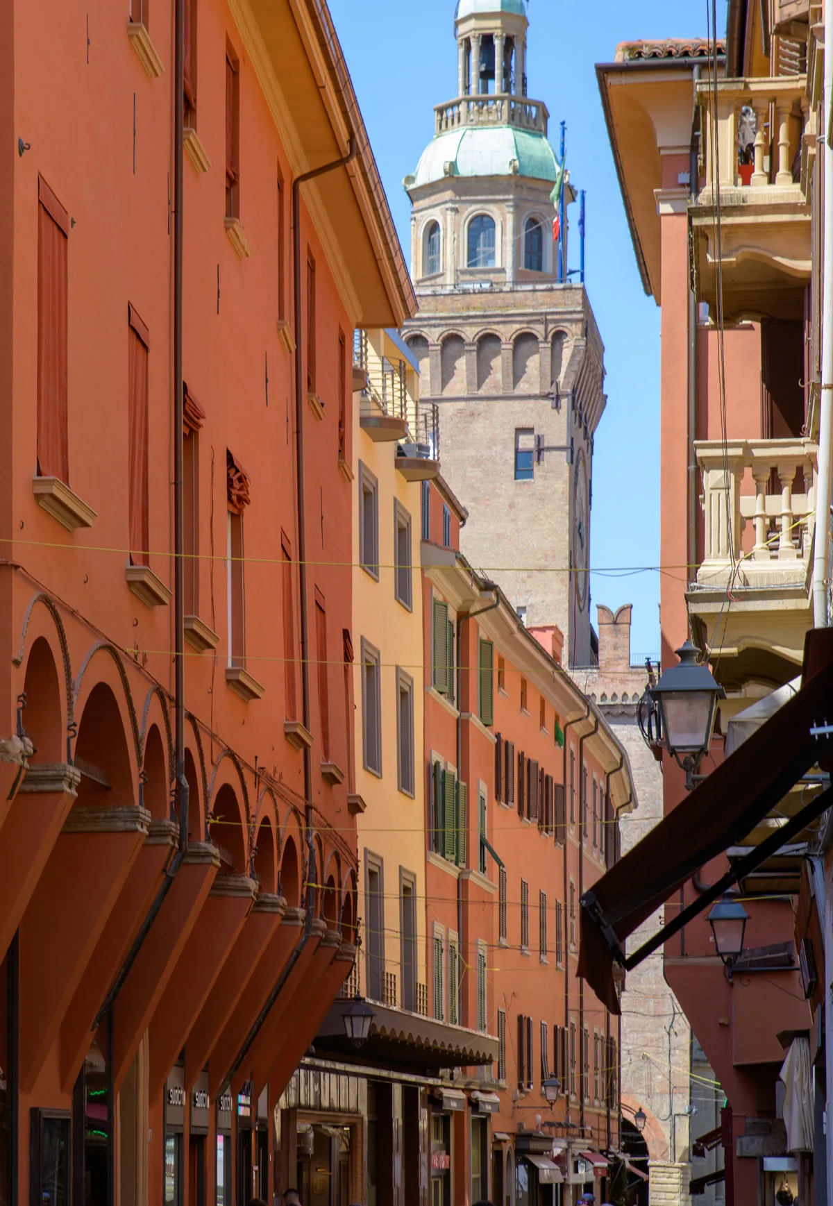 A narrow street with orange hued buildings in Bologna reveals a church tower at the end of the street. 