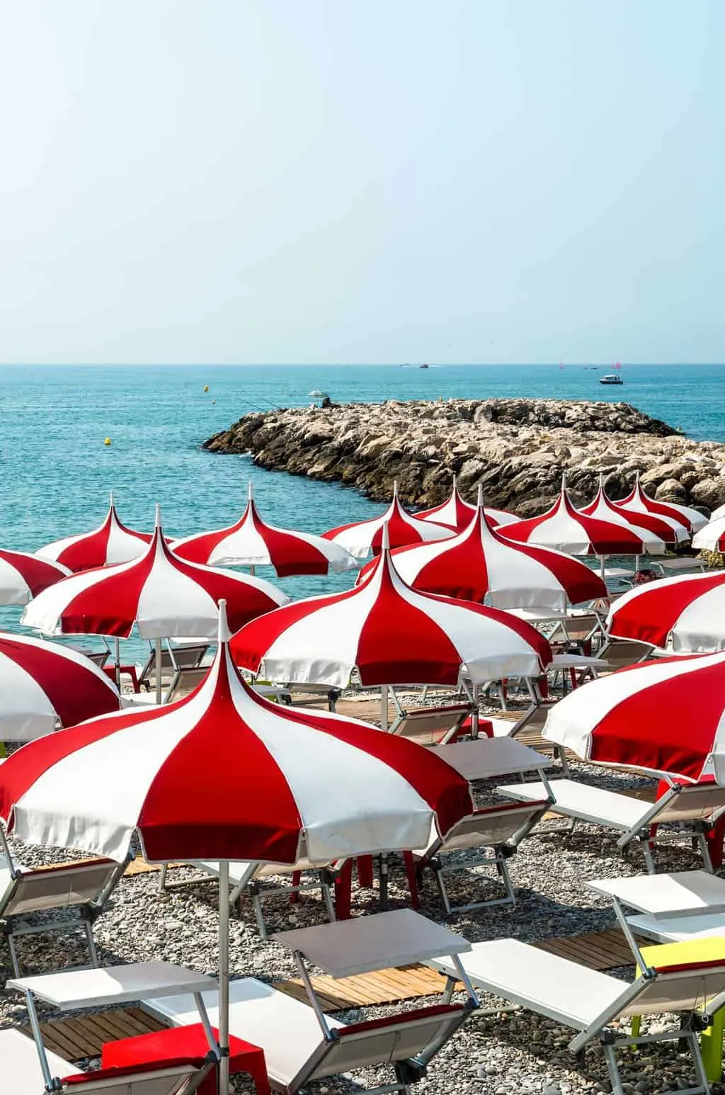 Red and white umbrellas and sun lounges line the pebble beach in Cagnes-sur-Mer overlooking turquoise blue water on a sunny day.