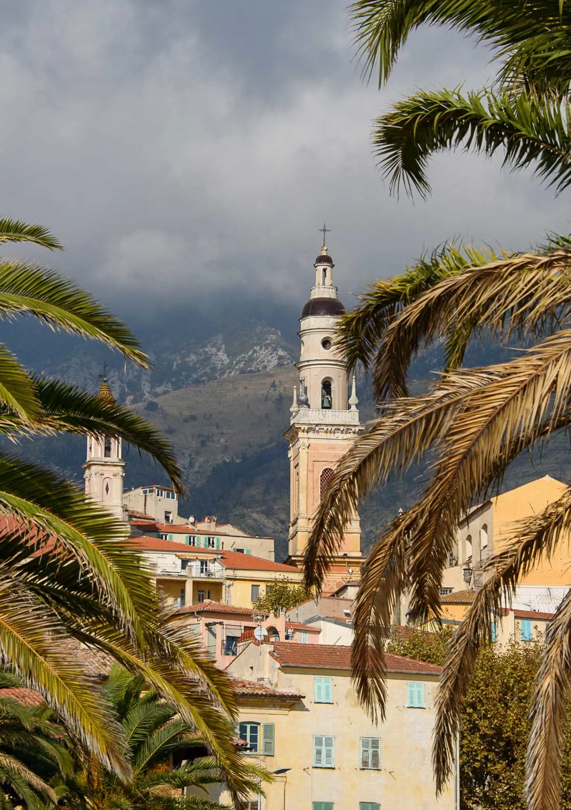 Pink hued village with baroque church spires in front of misty mountains with palm trees in the foreground. 