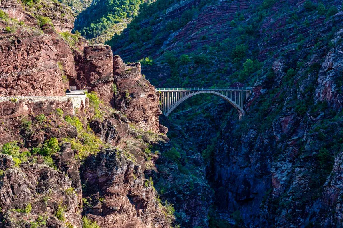 A bridge crosses a narrow gorge with steep red rocks and a winding road on one side. 